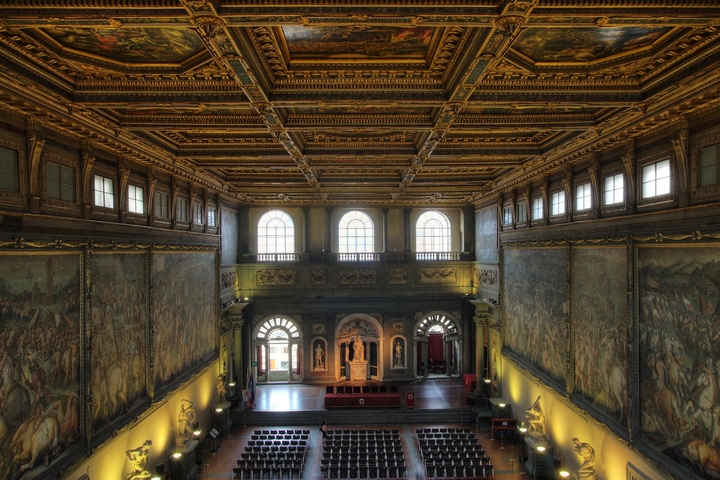Le Salone dei Cinquecento au Palazzo Vecchio. 54 mètres de long, 23 de large, 18 de haut. Au-dessus des tables du dîner, un immense plafond à caissons; sur les murs, les compositions monumentales réalisées sous la direction de Giorgio Vasari pour Côme Ier de Médicis, exaltant Florence, ses conquêtes et le pouvoir ducal.